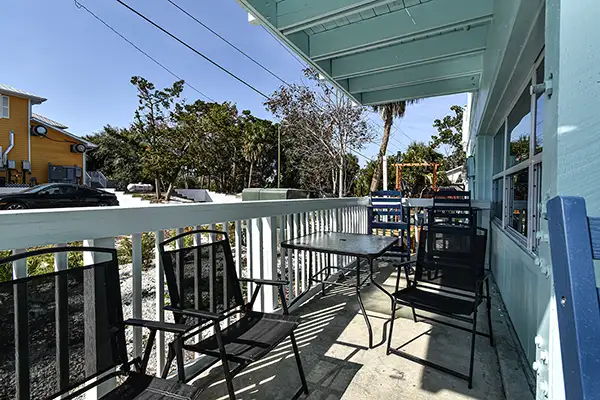 132-Balcony Turtle of Cedar Key - Ocean View Vacation Rentals Outdoor balcony with black metal chairs around a small table, turquoise building to the right, overlooking a sunlit street with trees and power lines.
