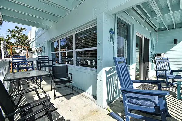 132-Porch Turntle of Cedar key - Ocean View Vacation Rentals Sunny outdoor patio on a turquoise house featuring a metal table, black and blue rocking chairs, and a sliding glass door nearby on a concrete floor.