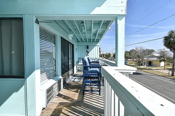 230-Back Porch Cedar Key Dophin Cay - Ocean View Vacation Rentals Turquoise motel balcony with white railing and blue outdoor chairs facing the street, under a covered walkway on a sunny day.