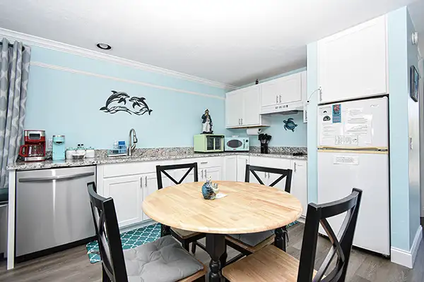 230-Dining room with beach decor - Ocean View Vacation Rentals Cozy small coastal kitchen with a round wooden table, four chairs, white cabinets, stainless appliances, blue walls with dolphin decals.
