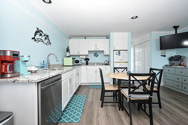 230-Kitchen Ocean Blue - Ocean View Vacation Rentals Coastal kitchen with white cabinets and turquoise walls, dolphin wall art, stainless appliances, and a round table with four black chairs.