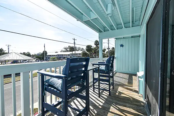230-Porch Overlooking beach - Ocean View Vacation Rentals Turquoise-paneled balcony with blue wooden chairs and a small table, overlooking a sunny street neighborhood below.